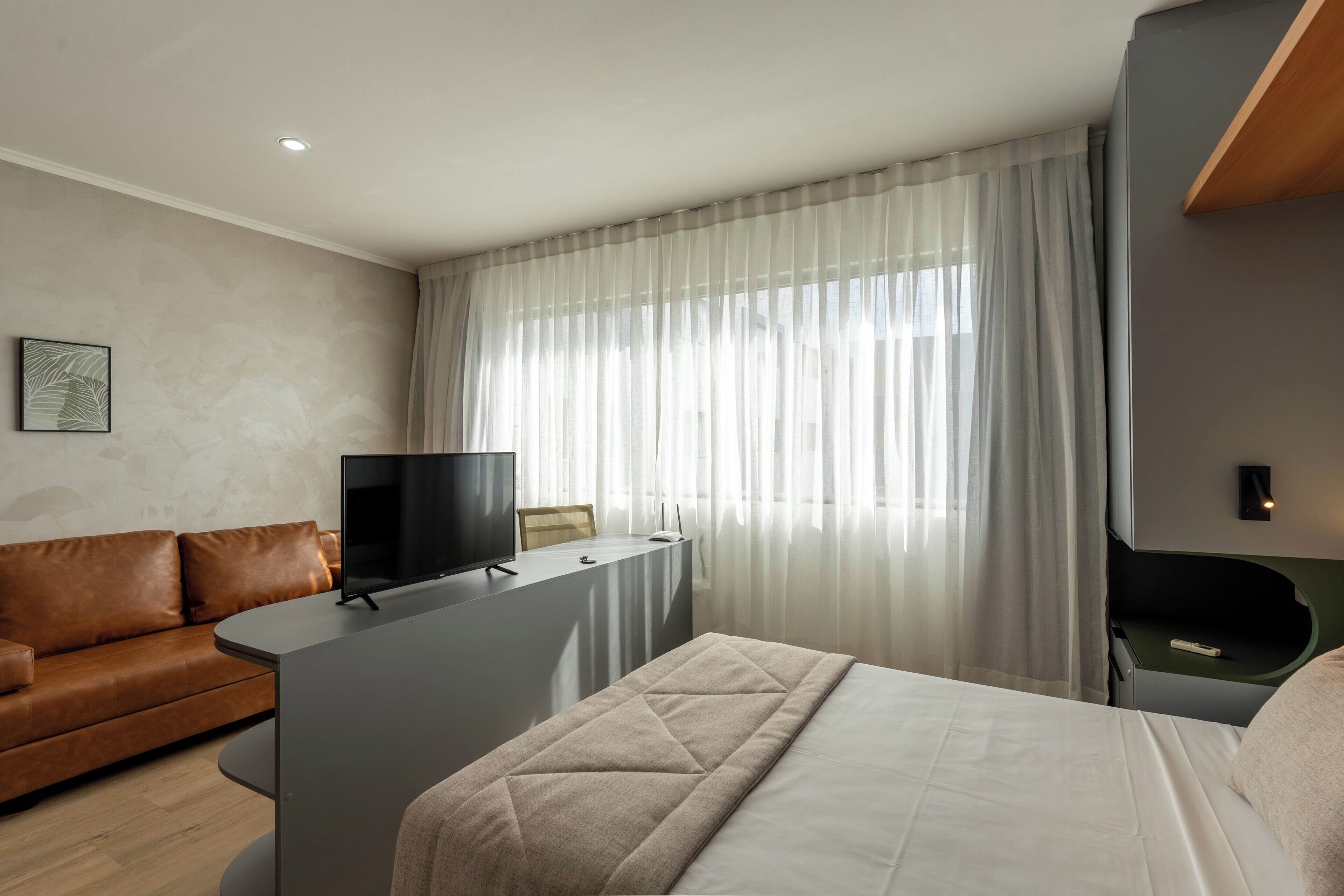 Ett modernt hotellrum med en säng, ett skrivbord med en tv, en lädersoffa och ett stort fönster med tunna, genomskinliga gardiner.