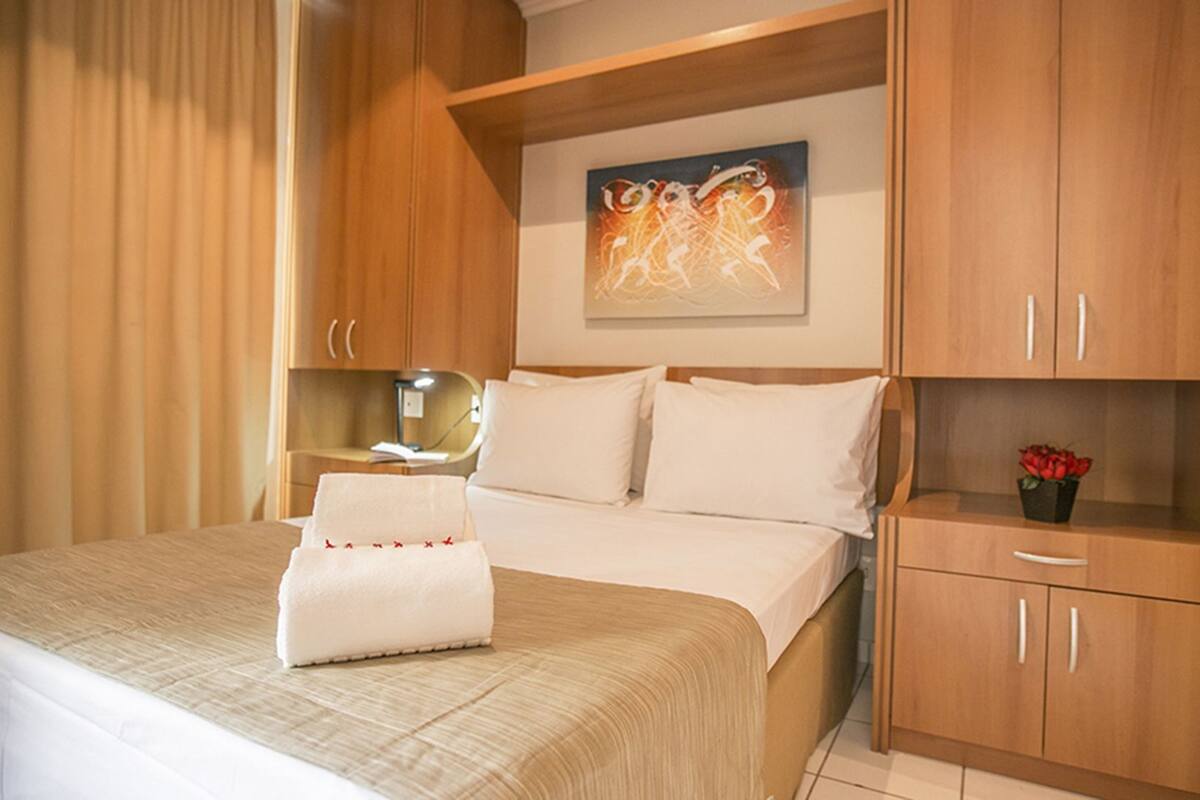 Une chambre d’hôtel avec un lit, une table de chevet, une lampe, un tableau au mur et une petite armoire contenant un vase de fleurs.