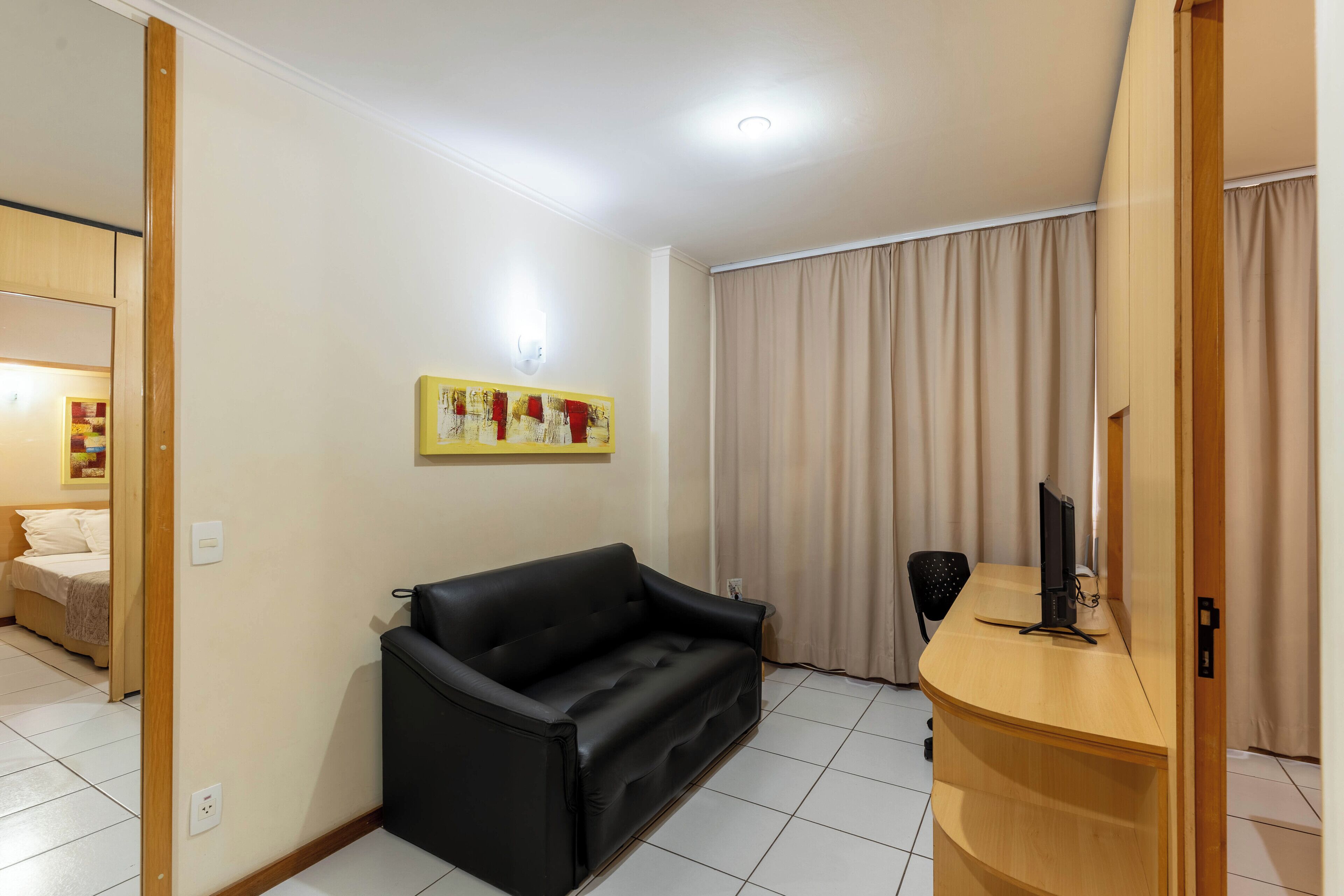 Ein Zimmer mit einer schwarzen Couch, einem Holztisch mit einem Computer, einem Stuhl, einem Bett mit weißem Kopfteil und einem beigefarbenen Vorhang.