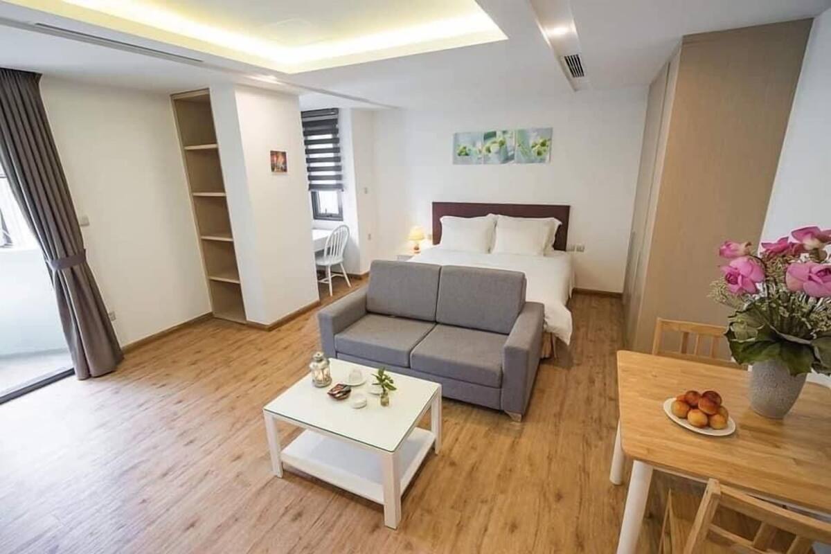 A modern hotel room with a sofa bed, a small table with a vase of flowers, and a dining table with a plate of fruit.