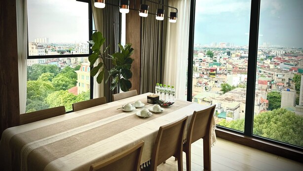 A modern dining room with a large table, chairs, and a view of a cityscape through large windows. - Ficus Suites Apartment (Hanoi)