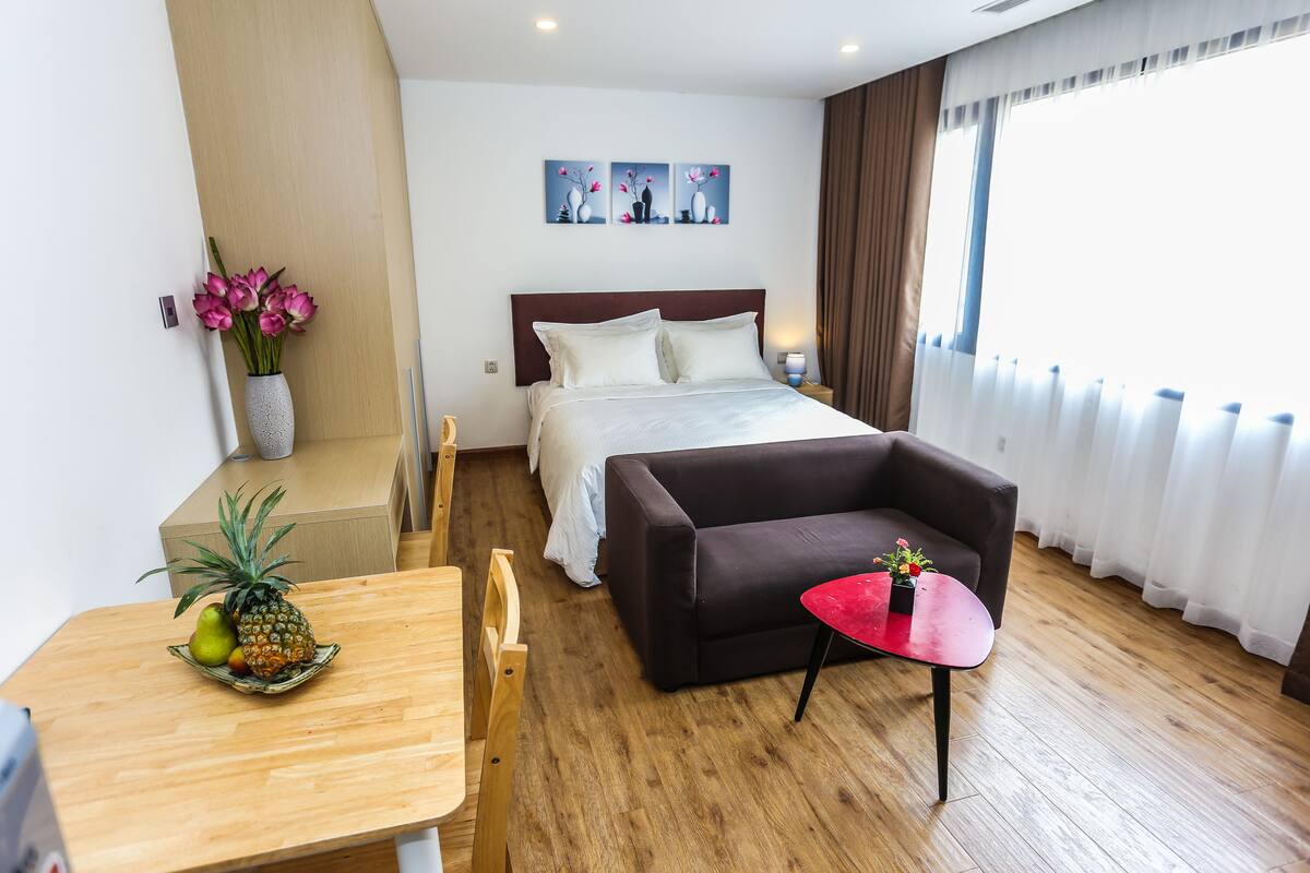 A hotel room with a bed, a sofa, a small table with a red top, a wooden table with fruits, and a vase with flowers.