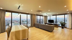 A spacious room with a dining table, sofa, and TV, large windows with a city view, and a ceiling fan. - Ficus Suites Apartment (Hanoi)