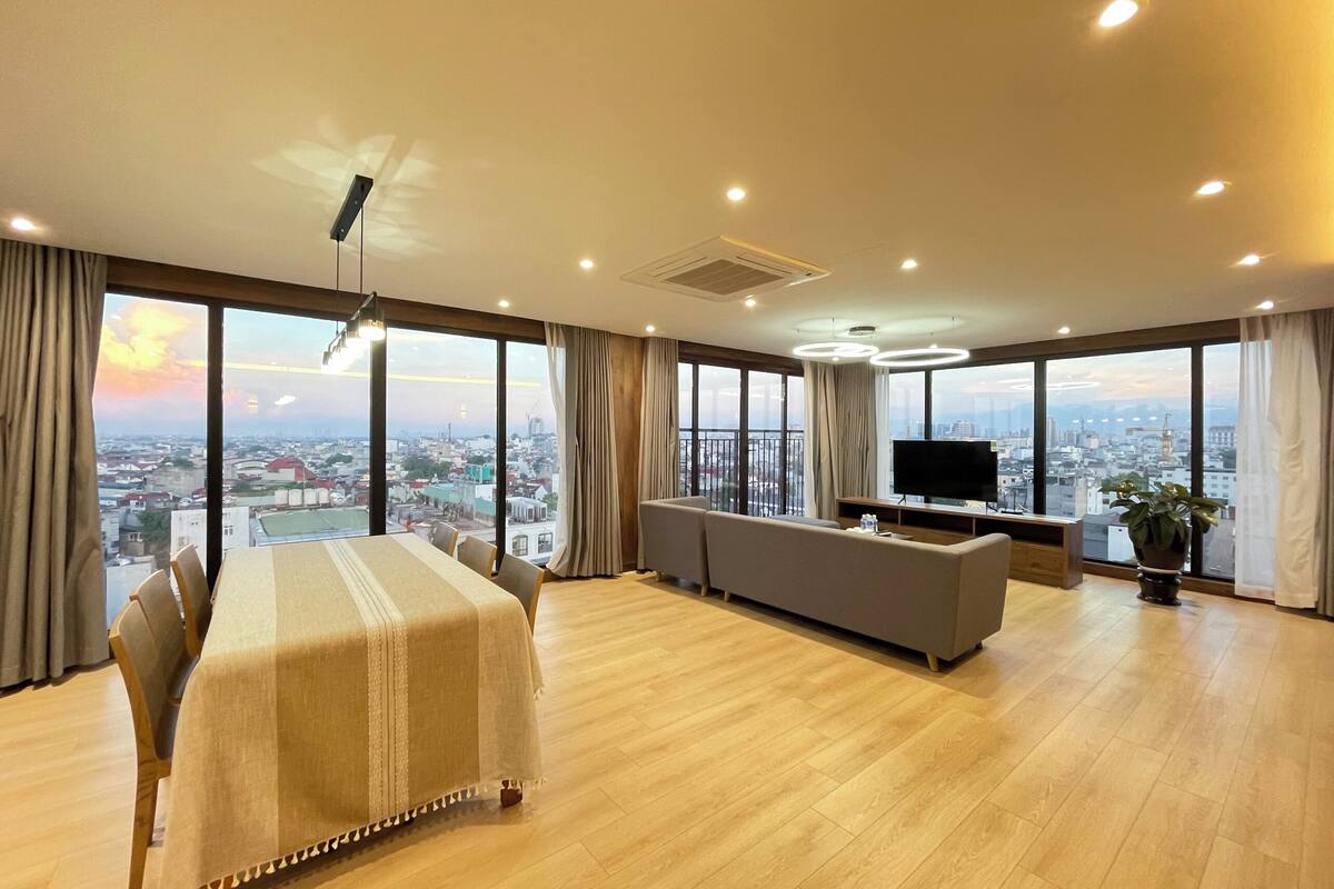 A spacious room with a dining table, sofa, and TV, large windows with a city view, and a ceiling fan.