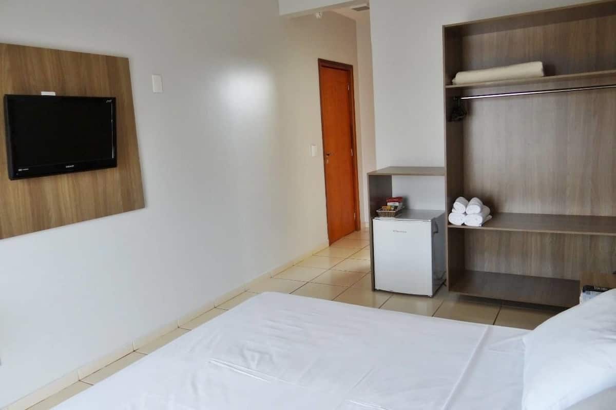 Ein Hotelzimmer mit einem Bett, einem an der Wand befestigten Fernseher, einem kleinen Kühlschrank und einem Holzboden mit Handtüchern.