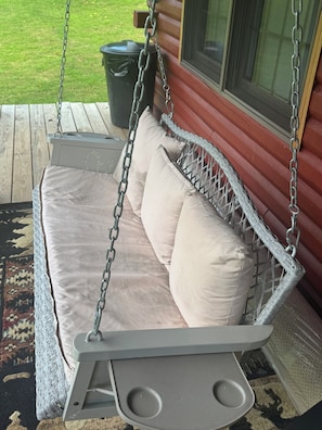 A white wicker swing bed with cushions, a small table, and a trash can nearby. - Log Cabin in the Field (Huntington)