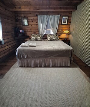 Un dormitorio de cabaña de madera con una cama, almohadas, una lámpara y una ventana con cortinas.