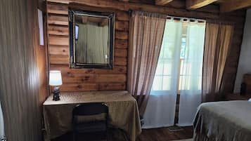 Una habitación con techo de madera, una mesa con lámpara, una silla, una cama y un ventanal con cortinas.