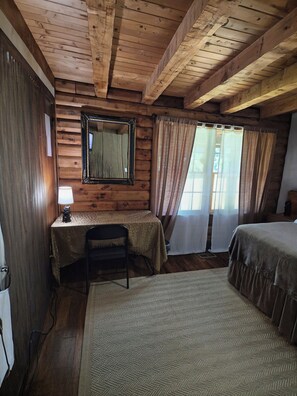 A room with a wooden ceiling, a table with a lamp, a chair, a bed, and a window with curtains. - Log Cabin in the Field (Huntington)