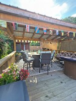 Un patio exterior con terraza de madera, mobiliario de mimbre y un área de estar cubierta. Banderines colgados arriba y un cartel de bienvenida.