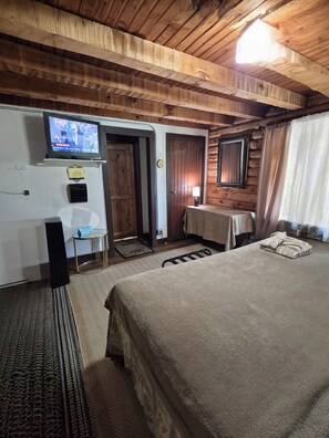 A wooden cabin-style room with a bed, a television mounted on the wall, a small table, and a door leading to another room.