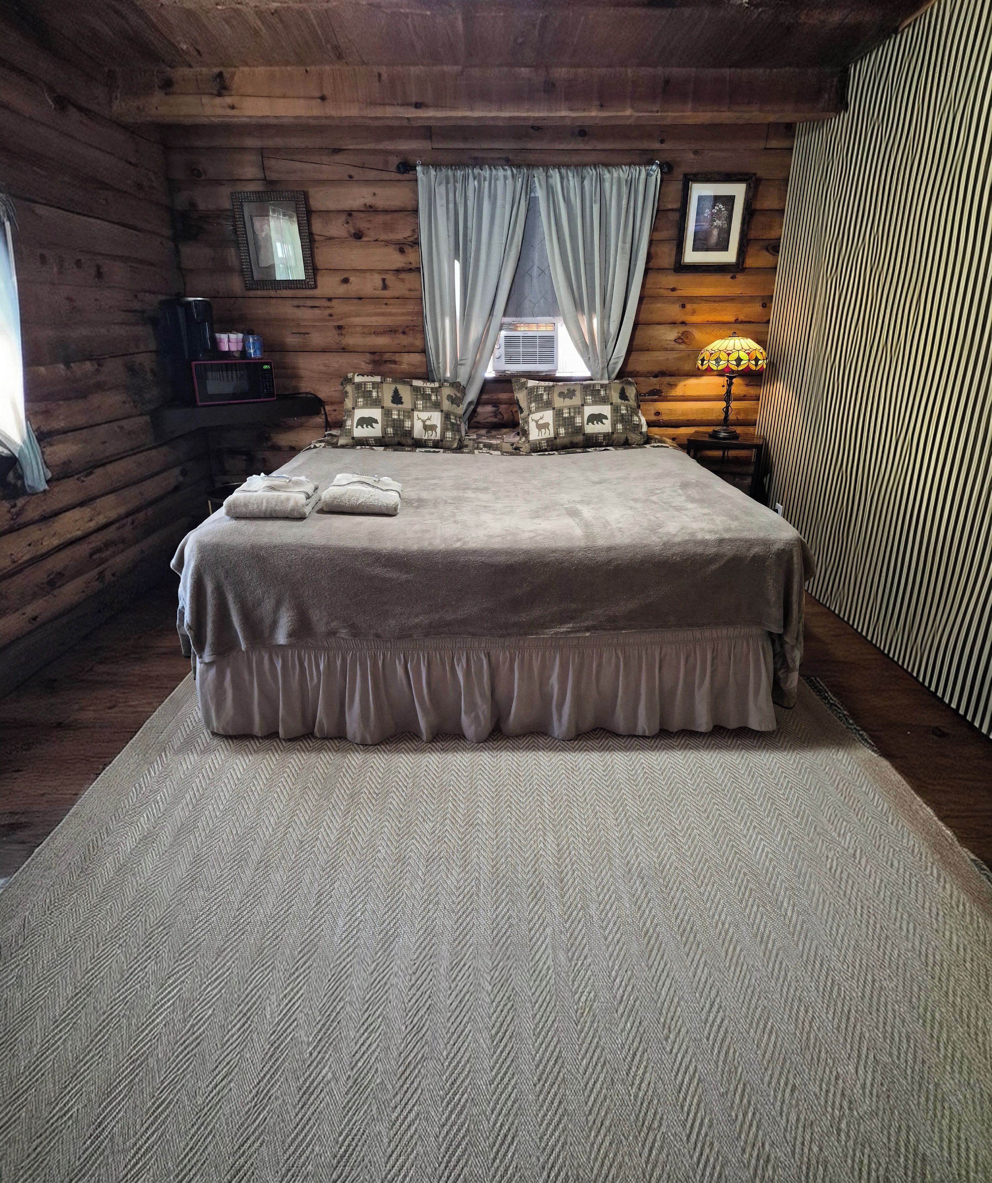 Un dormitorio de cabaña de madera con una cama, almohadas, una mesita de noche, una lámpara y una ventana con cortinas.