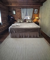 Un dormitorio de cabaña de madera con una cama, almohadas, una mesita de noche, una lámpara y una ventana con cortinas.