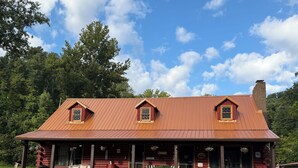 A log cabin with a red metal roof, two dormer windows, a porch with a bench, and a well-maintained lawn. - Log Cabin in the Field (Huntington)