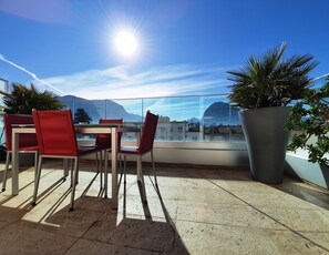 Una terraza con mesa y sillas, una planta grande en maceta y vistas a edificios y montañas.