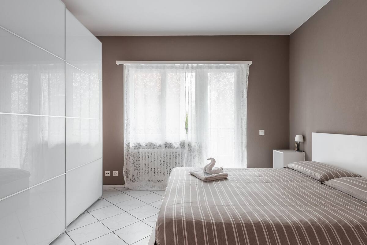Un dormitorio moderno con una cama, una mesita de noche, un ventanal con cortinas translúcidas y un armario blanco.