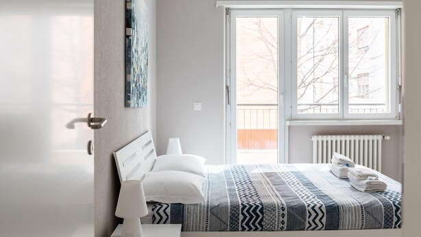 Un dormitorio con cama, mesita de noche, lámpara, ventana con vistas a árboles y radiador.