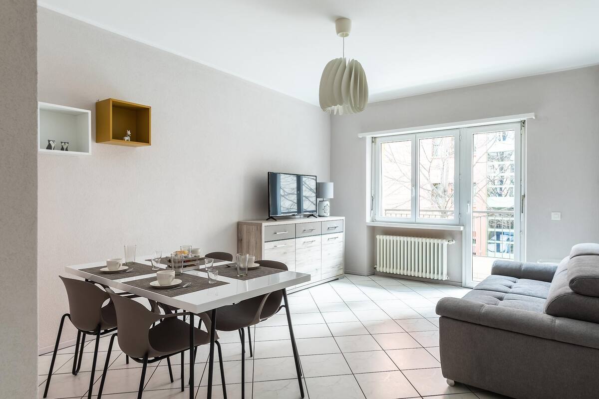 Una sala moderna con un sofá gris, una mesa de comedor con sillas, un televisor sobre un mueble de madera y un ventanal con vistas al exterior.