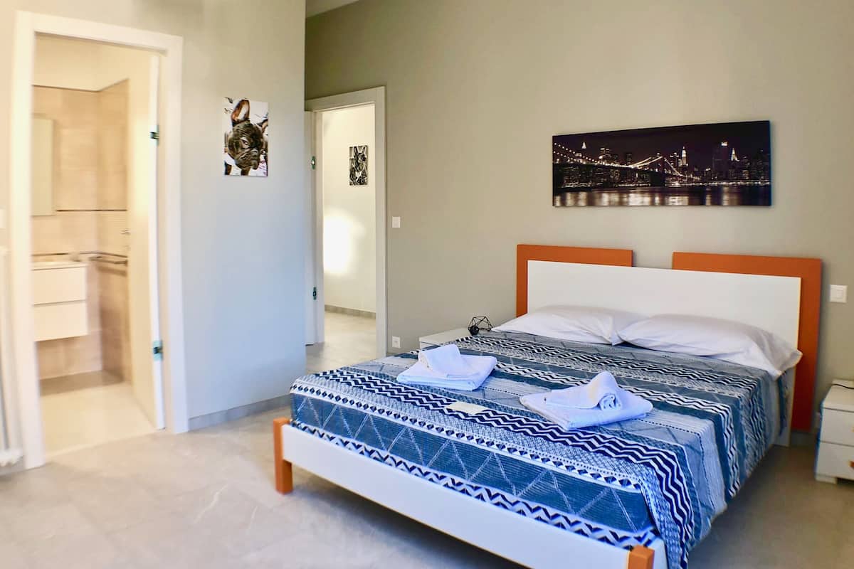 Un dormitorio con cama, mesita de noche, un cuadro colgado en la pared y una puerta que da al baño.