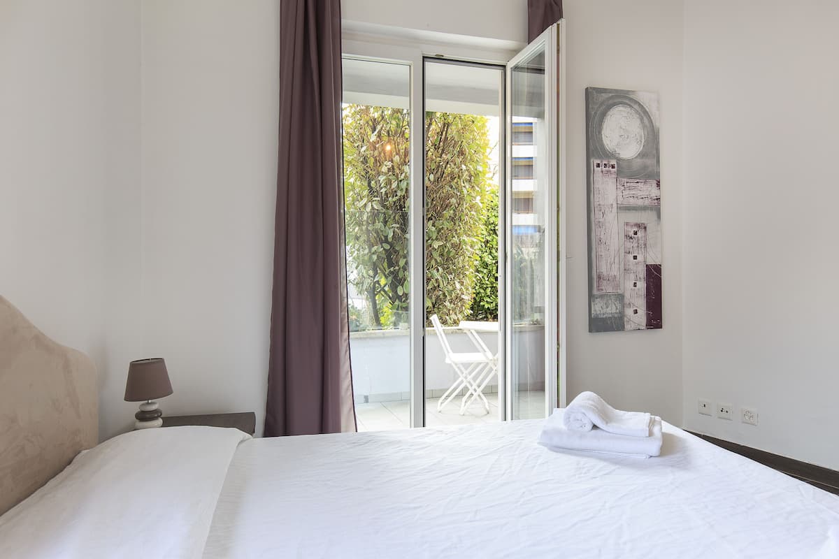 Habitación de hotel con cama, mesita de noche, lámpara, puerta corrediza de vidrio a un balcón y una obra de arte colgada en la pared.