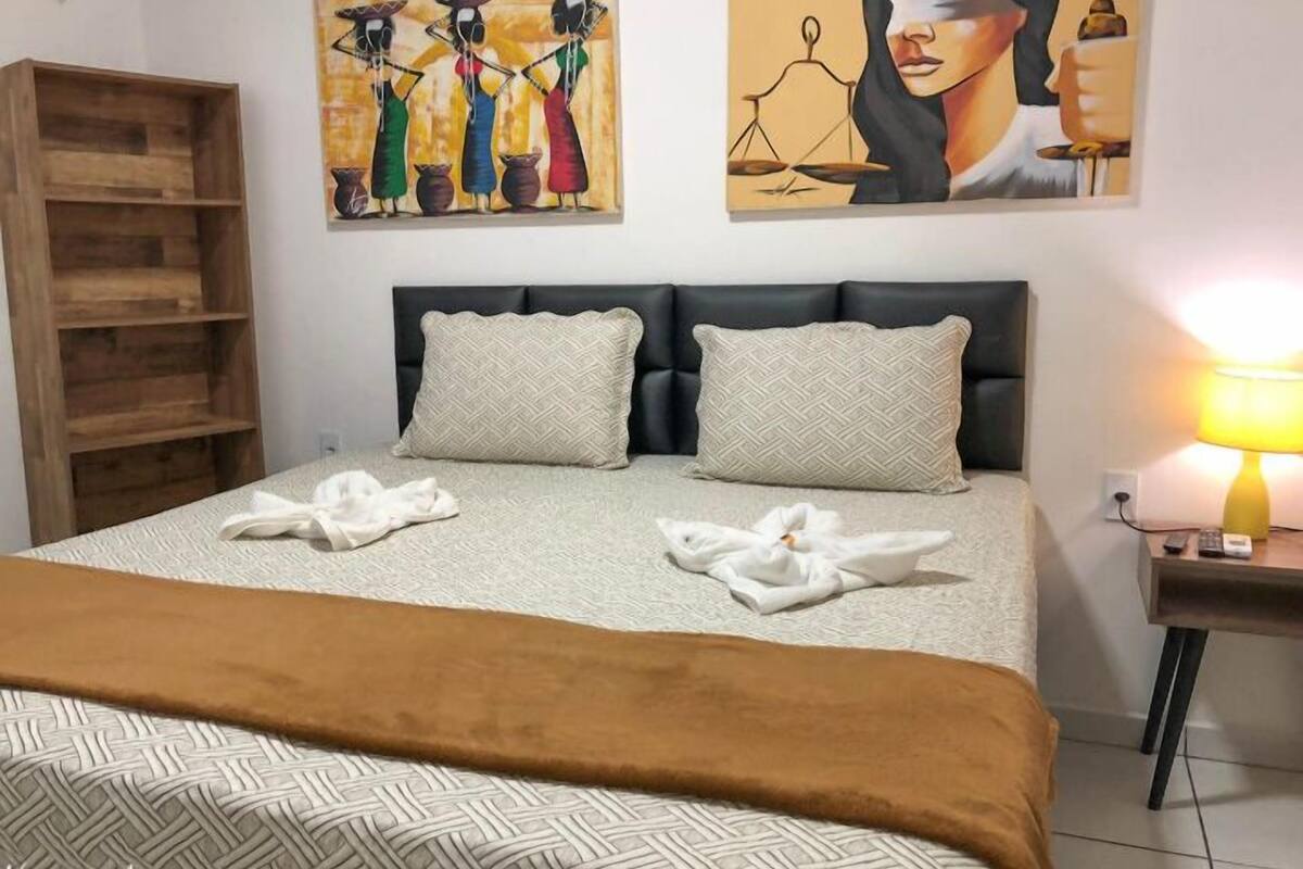 Una cama bien hecha con una manta marrón, dos almohadas y un cabecero. A la izquierda hay una estantería de madera y, a la derecha, una lámpara sobre una mesita.