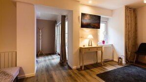 A room with a wooden floor, a desk with a lamp, a painting on the wall, a radiator, and a chair.