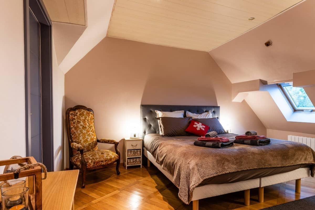 A cozy attic bedroom with a sloped ceiling, a large bed with a headboard, a chair, a nightstand, and a small table.