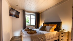 A neatly made bed with a headboard, a bedside table with drawers, a wall-mounted TV, and a window with curtains.