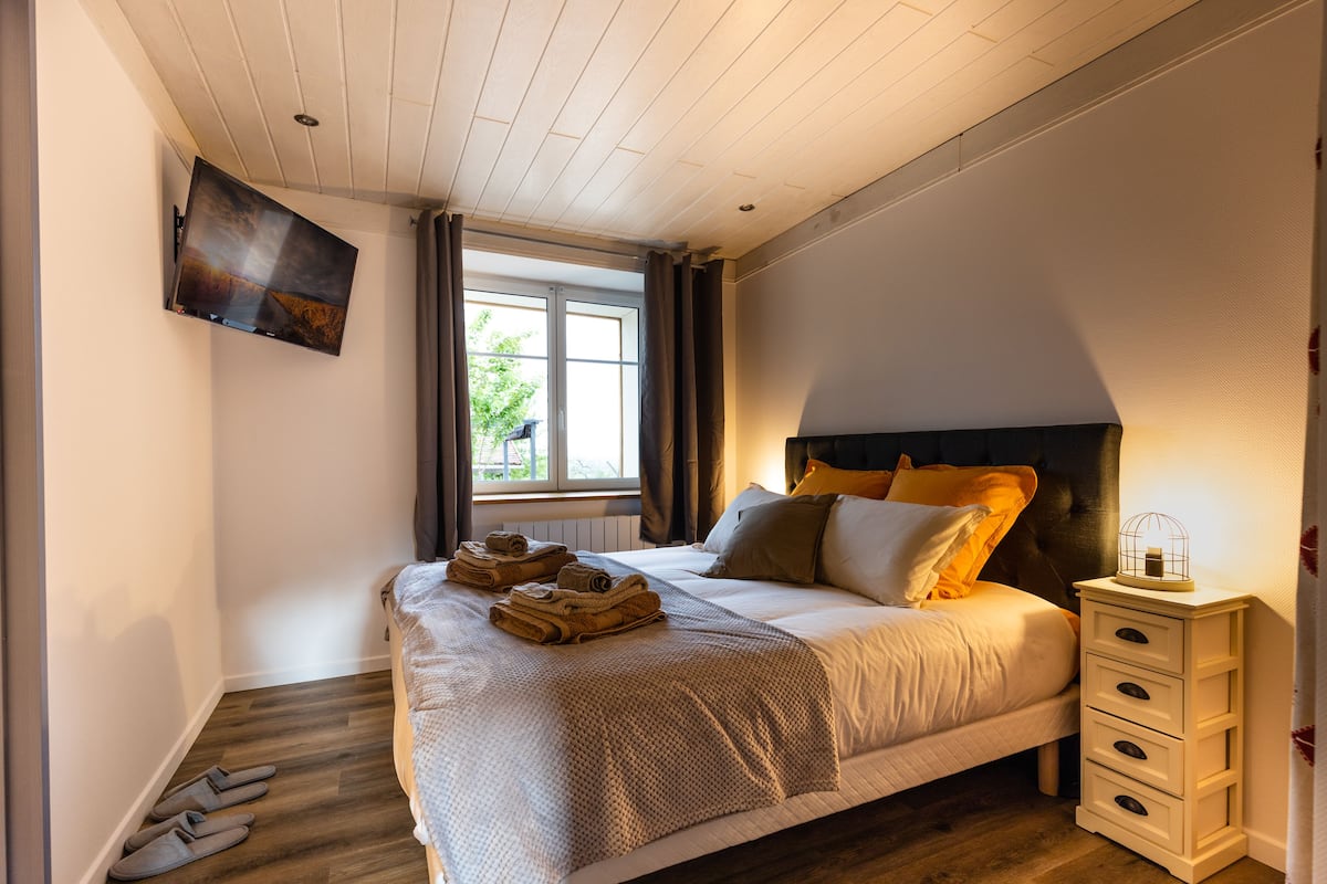 A neatly made bed with a headboard, a bedside table with drawers, a wall-mounted TV, and a window with curtains.