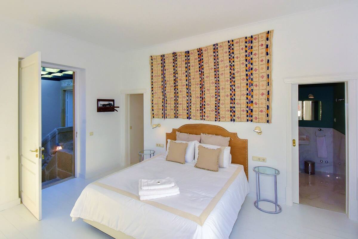 A bedroom with a bed, a headboard, a wall tapestry, a bedside table, and a bathroom door.