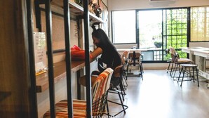 Un café con una decoración moderna, que presenta una barra larga con sillas, una persona atendiendo detrás de la barra y grandes ventanales que proporcionan luz natural.