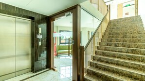 Un hall d’hôtel moderne avec un sol en marbre, un ascenseur en verre et un escalier menant à un étage supérieur.