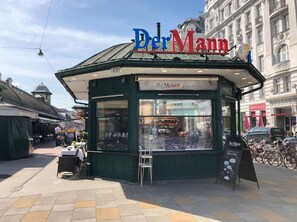 A small, green-roofed kiosk with 'Der Mann' signage, located on a pedestrian street with buildings and bicycles in the background.