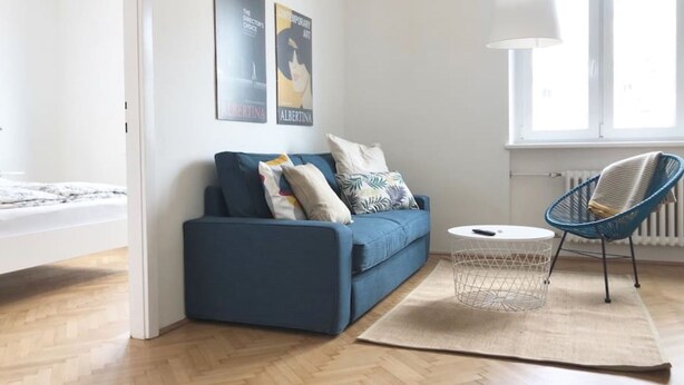 A modern living room with a blue sofa, a round coffee table, and a blue chair. There are two posters on the wall and a window providing natural light. - Lovely Apartment directly at Naschmarkt (Vienna)