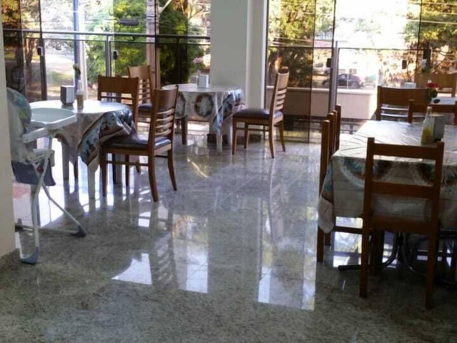 A dining area with a polished granite floor, wooden chairs, and tables. Large windows provide views of trees and a road outside.