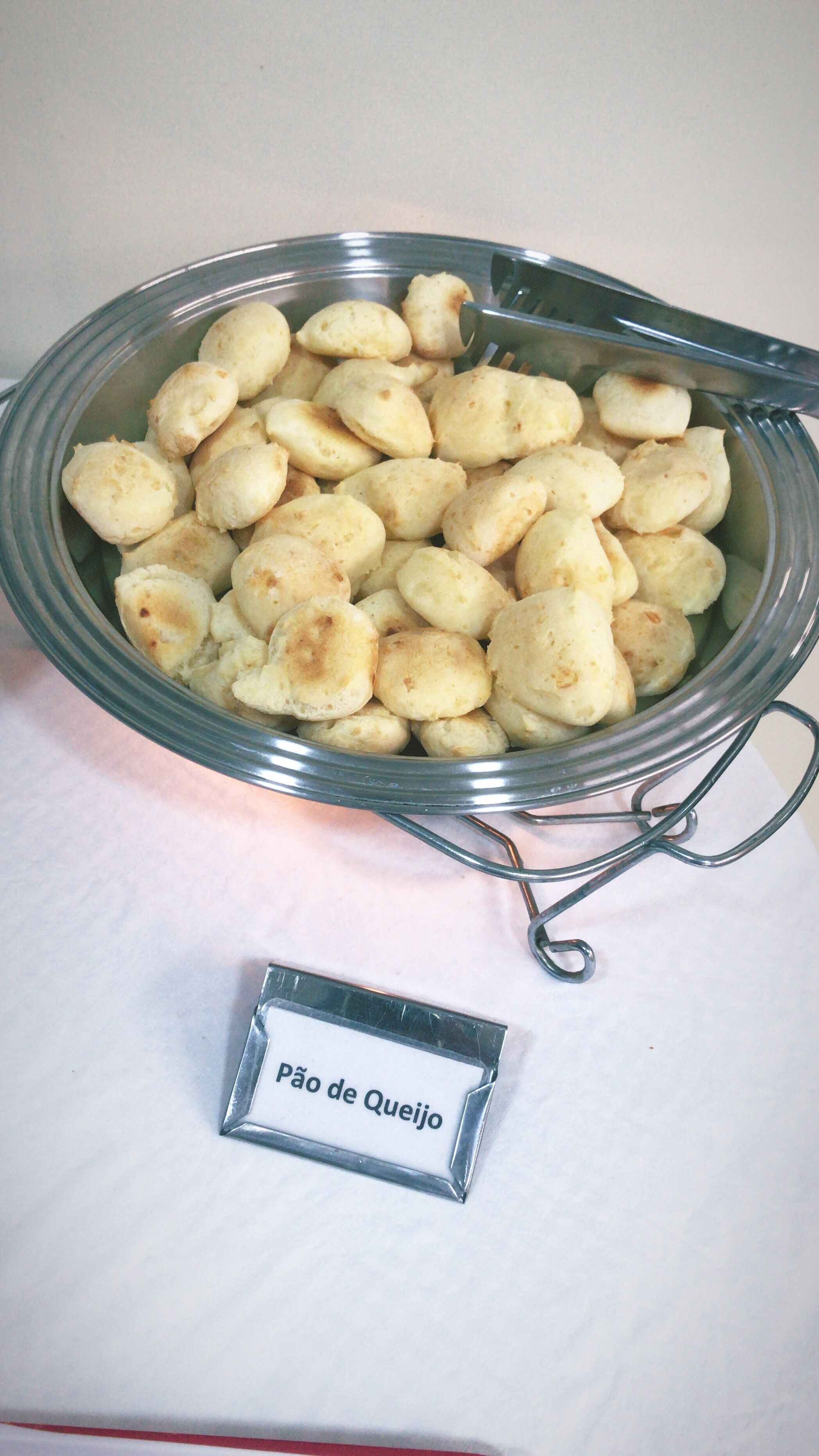 Un cuenco de vidrio lleno de pão de queijo, un tipo de pan de queso brasileño, con una cuchara de metal en su interior. Una pequeña etiqueta de metal con la inscripción 'Pão de Queijo' está colocada junto al cuenco.