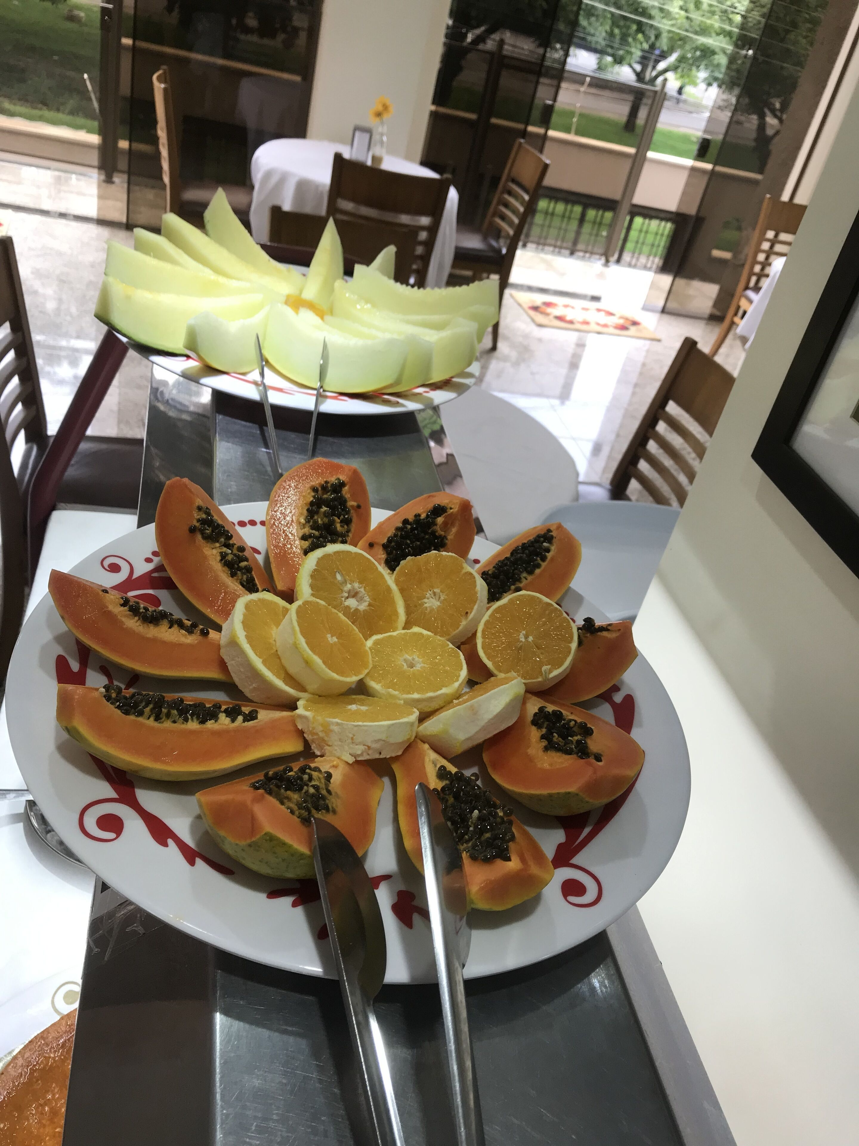 Une table sur laquelle est disposée une assiette de fruits comprenant de la papaye et des oranges. On aperçoit une porte vitrée et des sièges d’extérieur à l’arrière-plan.