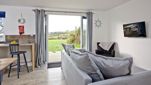 A modern living room with a grey sofa, a dining table, and a kitchen area with a view of the countryside. - Wheal Dream (Helston)