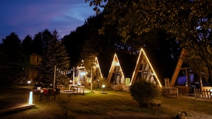 Un terrain de camping doté de cabanes en forme de « A » en bois, d’un espace extérieur aménagé pour s’asseoir et de guirlandes lumineuses.