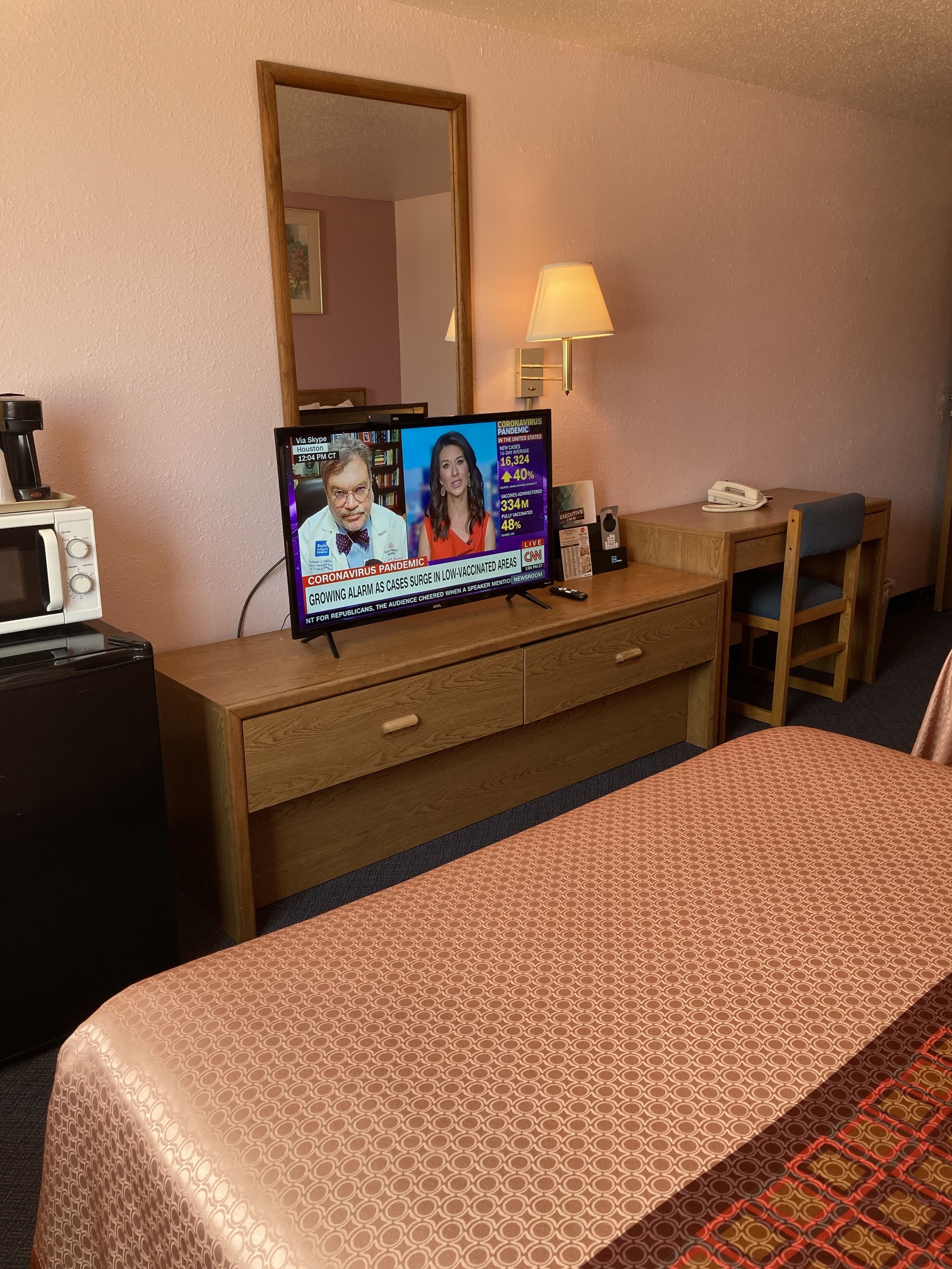 A hotel room with a bed, a wooden desk, a television displaying a news broadcast, a telephone, and a lamp.