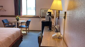 A hotel room with two beds, a desk with a lamp, a chair, a small table with a flower, and a window with curtains.
