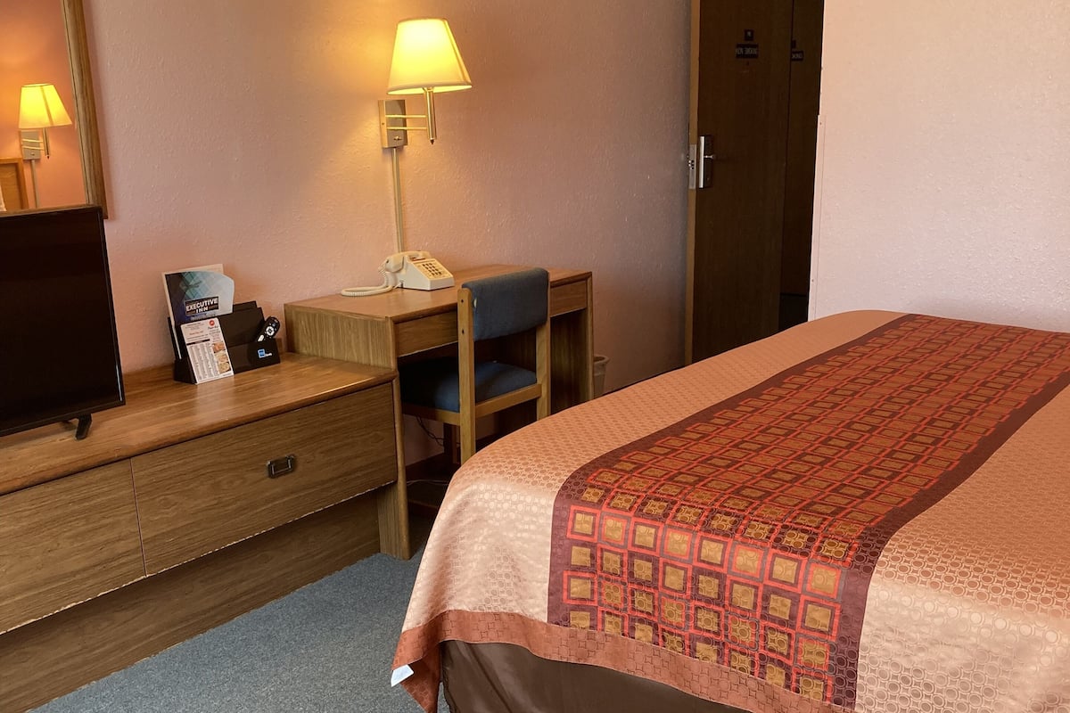 A hotel room with a bed, a desk with a lamp, a chair, a dresser, and a mirror.
