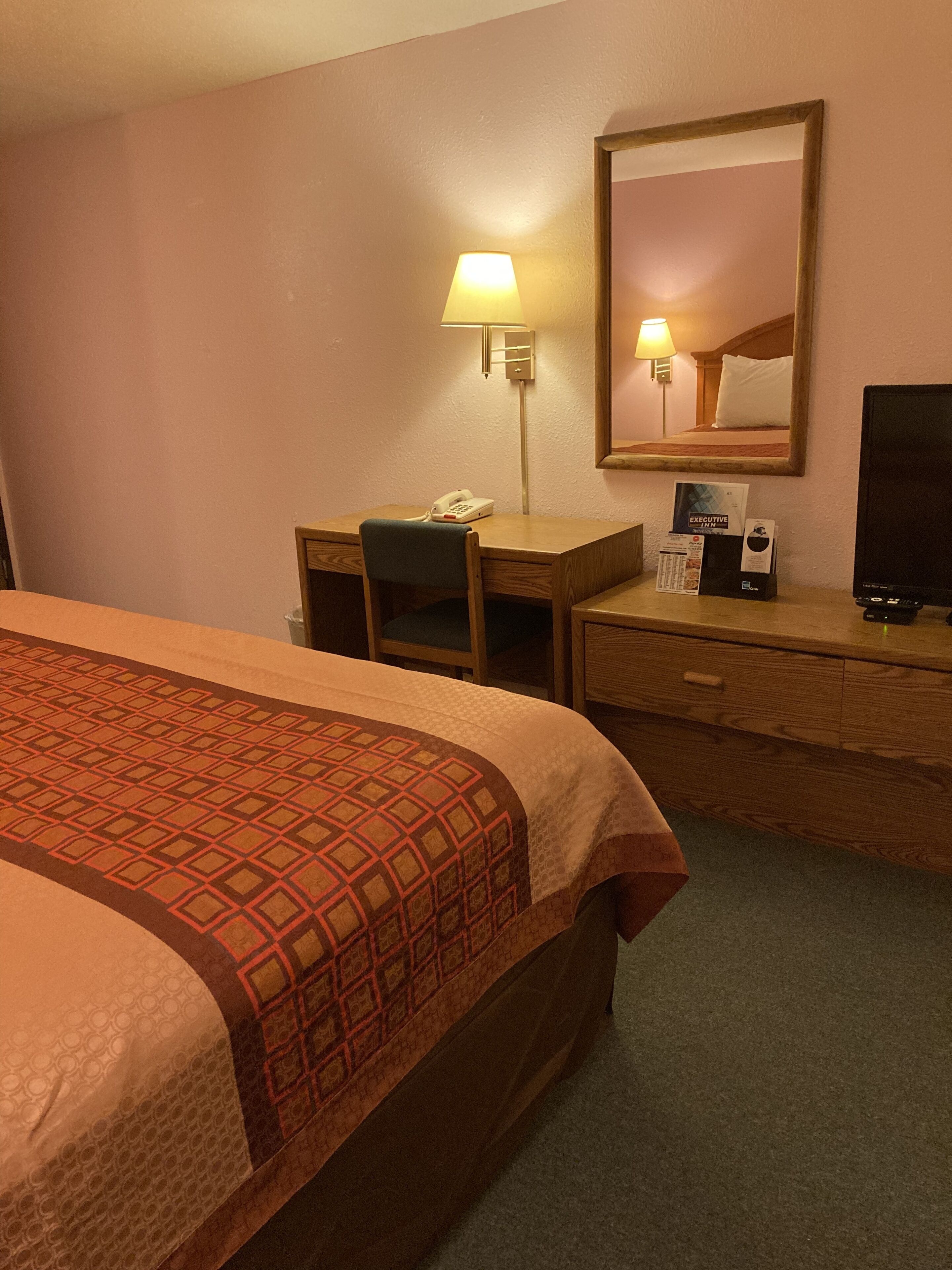 A hotel room with a bed, a desk with a telephone, a lamp, a mirror, and a television.