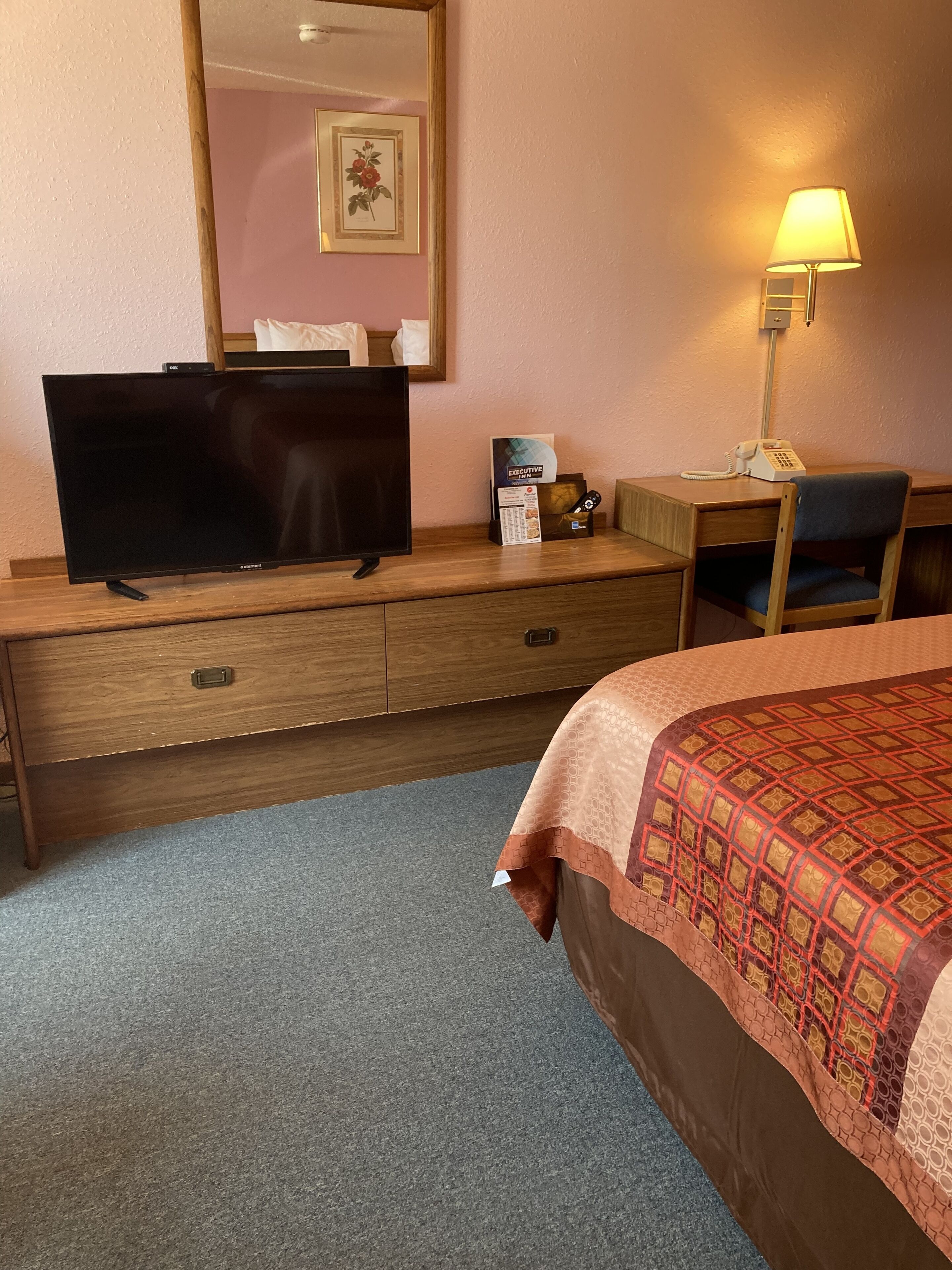 Habitación de hotel con un escritorio de madera, televisión, teléfono, una silla y una cama con edredón estampado.
