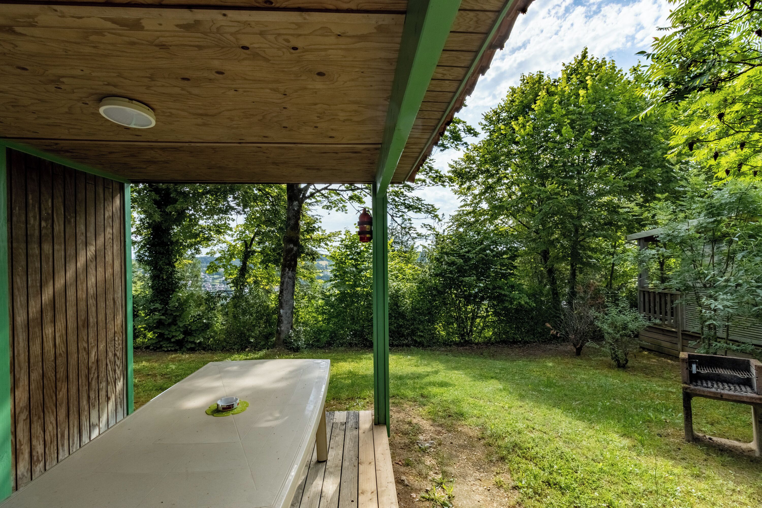 Een overdekt terras met een tafel en bankje, omringd door een gazon en bomen.
