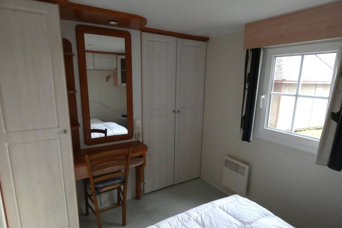 Une chambre à coucher avec un lit, une chaise en bois, un grand miroir, une armoire et une fenêtre.