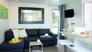 Een moderne woonkamer met een donkerblauwe bank, een klein bijzettafeltje, een flatscreen televisie aan de muur en een eethoek met een witte tafel en stoelen.