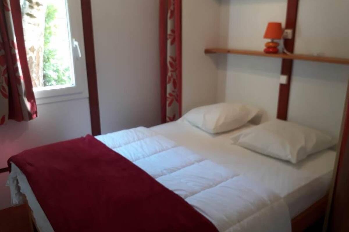 Un lit simple avec une literie blanche et une couverture rouge, une petite étagère avec une lampe et une fenêtre avec des rideaux.