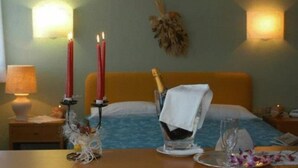 Habitació d'hotel amb un llit, una tauleta de nit amb espelmes, una ampolla de xampany i un vas amb flors.