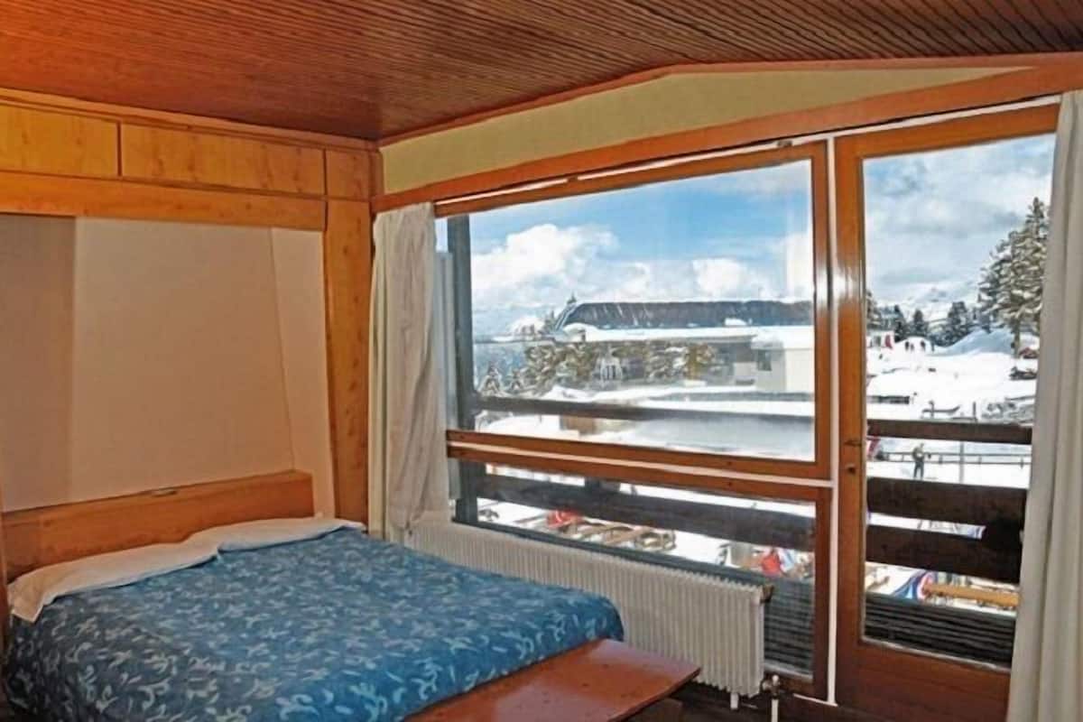 Une chambre à coucher avec un lit, une tête de lit en bois, une fenêtre donnant sur un paysage enneigé et un bâtiment.